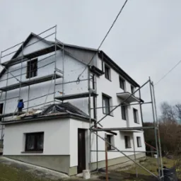 Dom w trakcie ocieplania z rusztowaniem: biała elewacja, styropian na ścianach, okna z brązowymi ramami. Częściowo ocieplona elewacja i pomalowana.