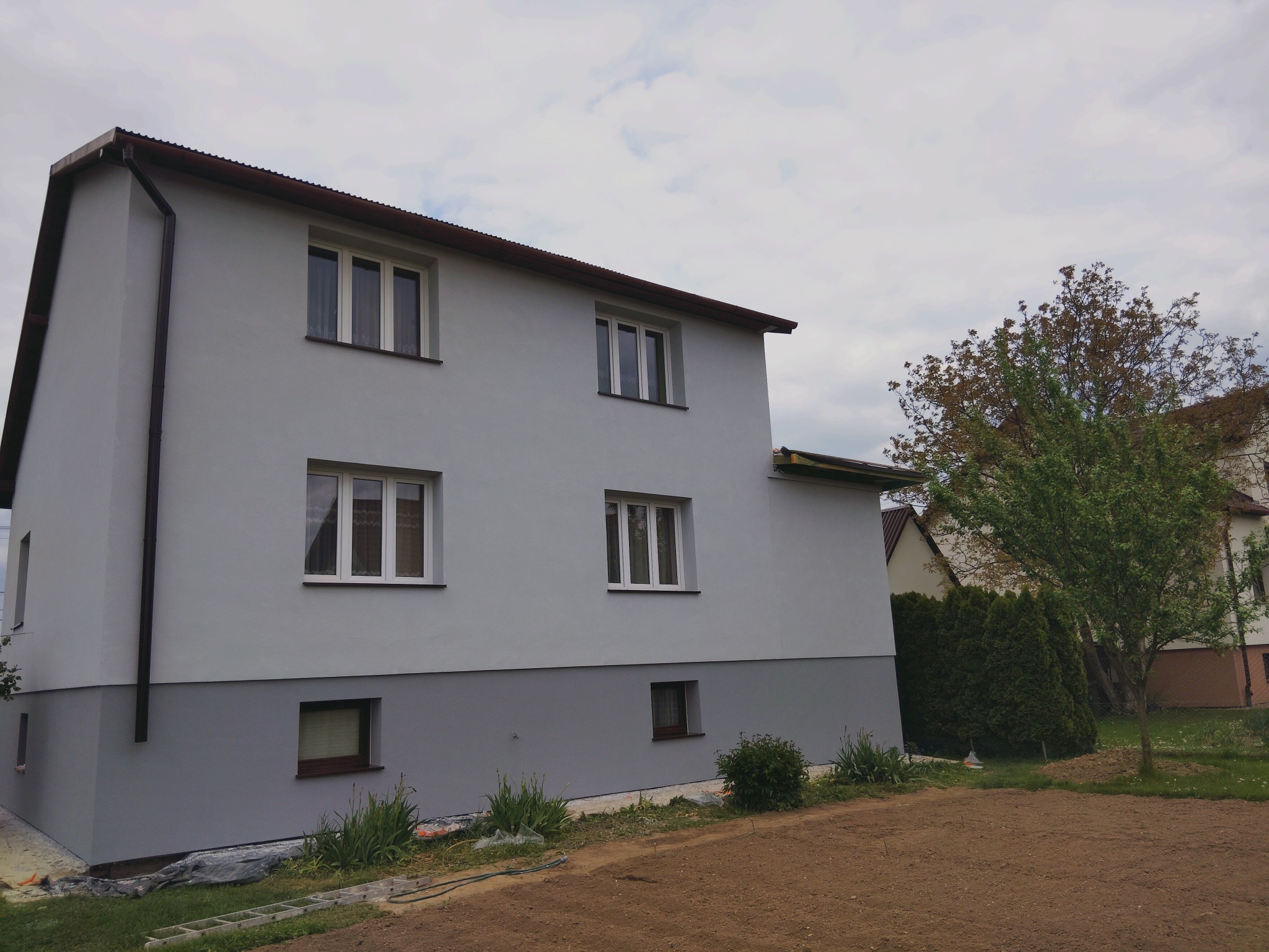 Dwukondygnacyjny dom po renowacji elewacji w odcieniach szarości, z widoczną dolną partią w ciemniejszym kolorze. W tle drzewa i fragmenty innych budynków.