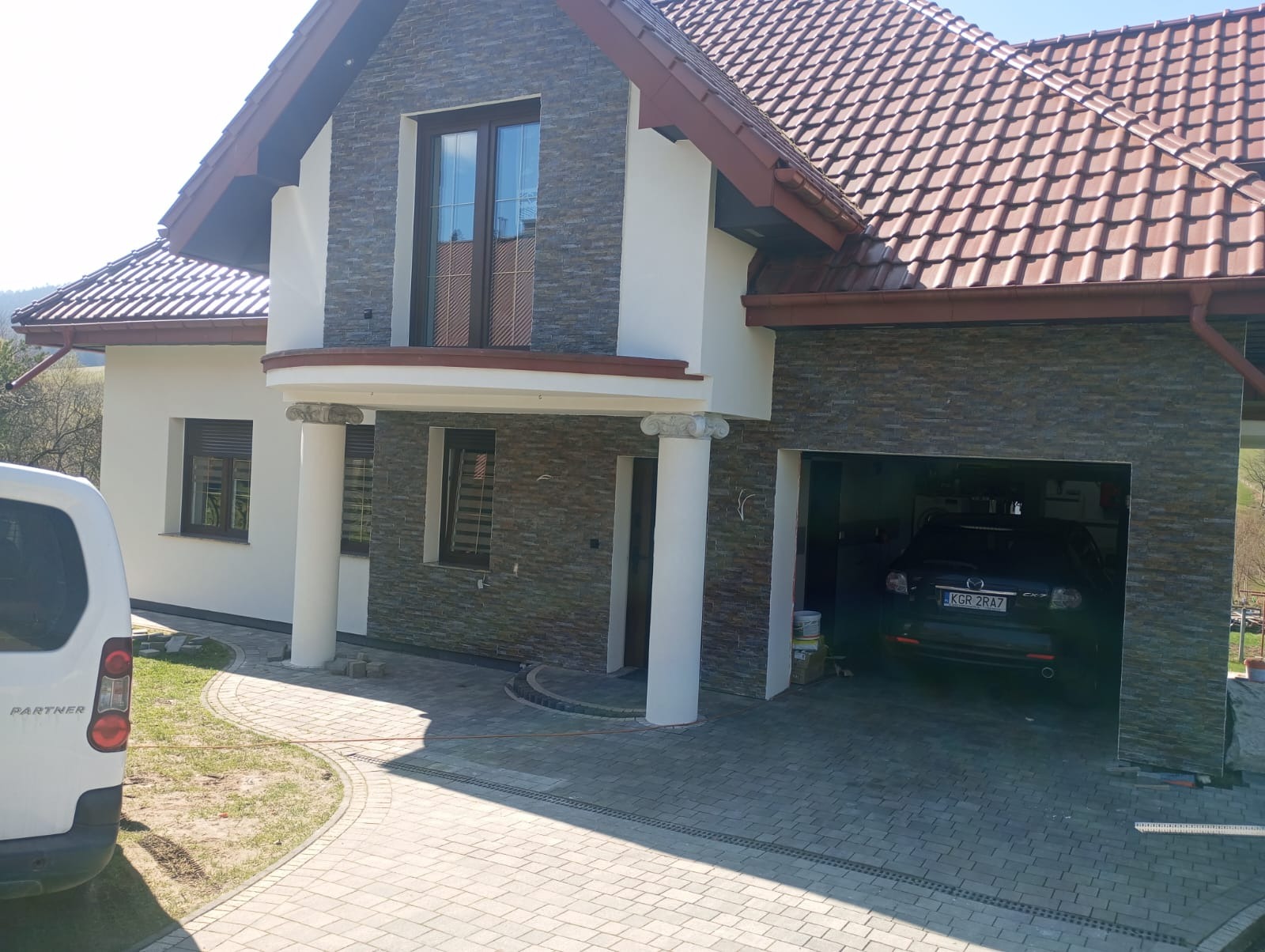 Dwukondygnacyjny dom z garażem, wykończony kamieniem elewacyjnym i białym tynkiem. Widoczny samochód w garażu i fragment chodnika z kostki brukowej. Częściowo widoczny samochód dostawczy.
