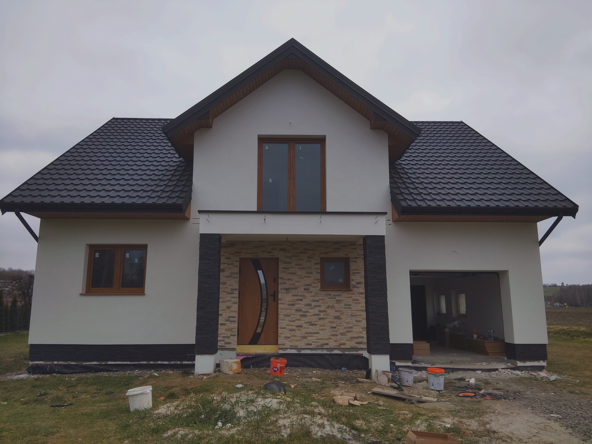 Nowoczesny dom jednorodzinny z elewacją w odcieniach bieli i szarości, z ciemnym dachem i drewnianą stolarką okienną, w trakcie prac wykończeniowych.