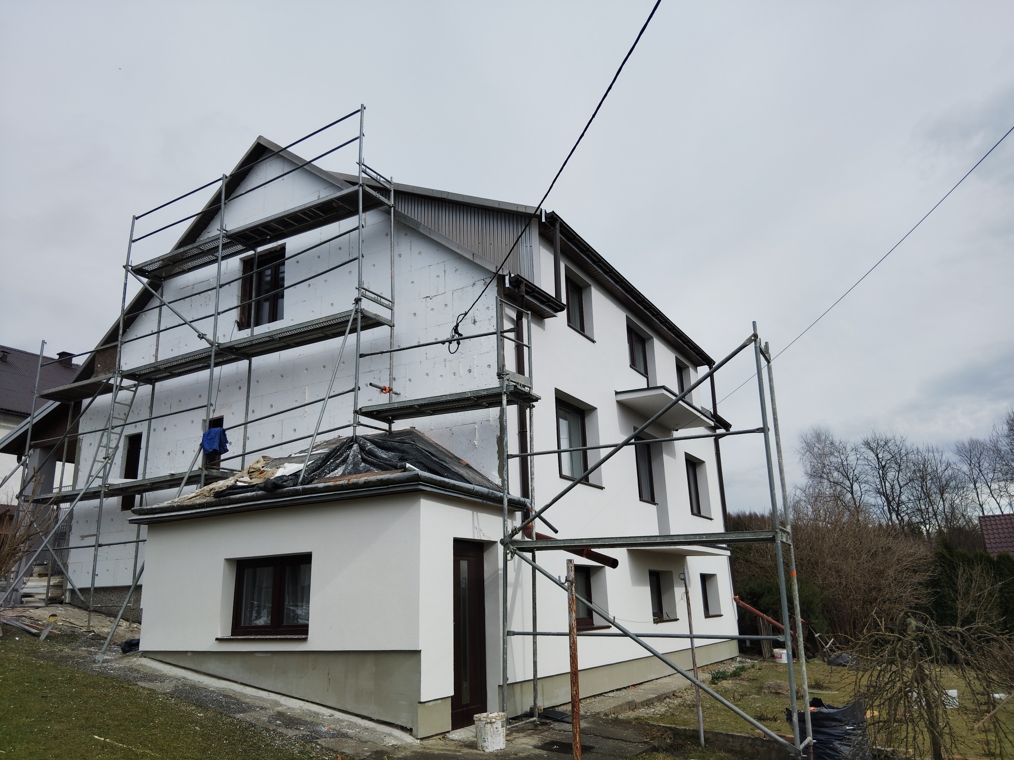 Dom w trakcie ocieplania z rusztowaniem: biała elewacja, styropian na ścianach, okna z brązowymi ramami. Częściowo ocieplona elewacja i pomalowana.