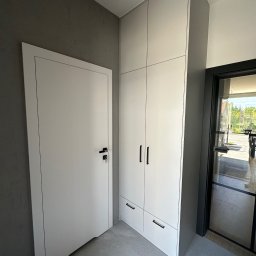 Ciach-Bud Rafał Jasieńczuk - Białe drzwi z czarną klamką sąsiadują z wysoką, jasną szafą wnękową z ciemnymi uchwytami na tle szarej ściany i podłogi z płytek. W tle widoczne przeszklone drzwi.