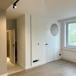 Ciach-Bud Rafał Jasieńczuk - Minimalistyczne wnętrze po remoncie: białe ściany z czarnymi akcentami, ukryte drzwi z szarym okręgiem, panele podłogowe i oświetlenie szynowe.