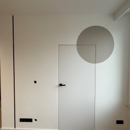 Ciach-Bud Rafał Jasieńczuk - Minimalistyczne wnętrze: biała ściana z ukrytymi drzwiami z czarną klamką, szary okrąg na ścianie, drewniana podłoga, czarne elementy dekoracyjne.