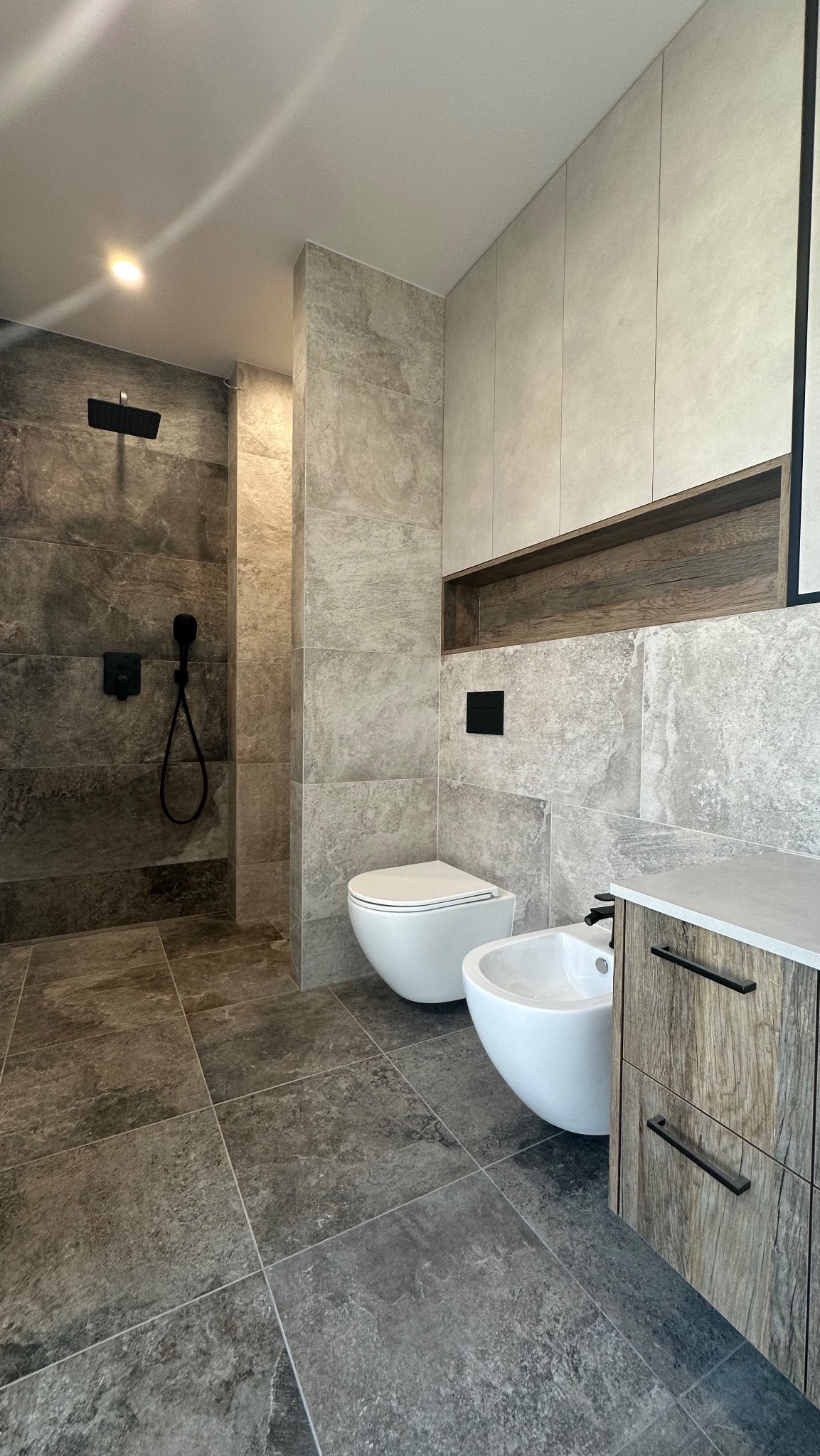 Nowoczesna łazienka z szarymi płytkami imitującymi kamień, kabiną prysznicową typu walk-in, podwieszaną toaletą i bidetem oraz szafką z drewnianym wzorem.