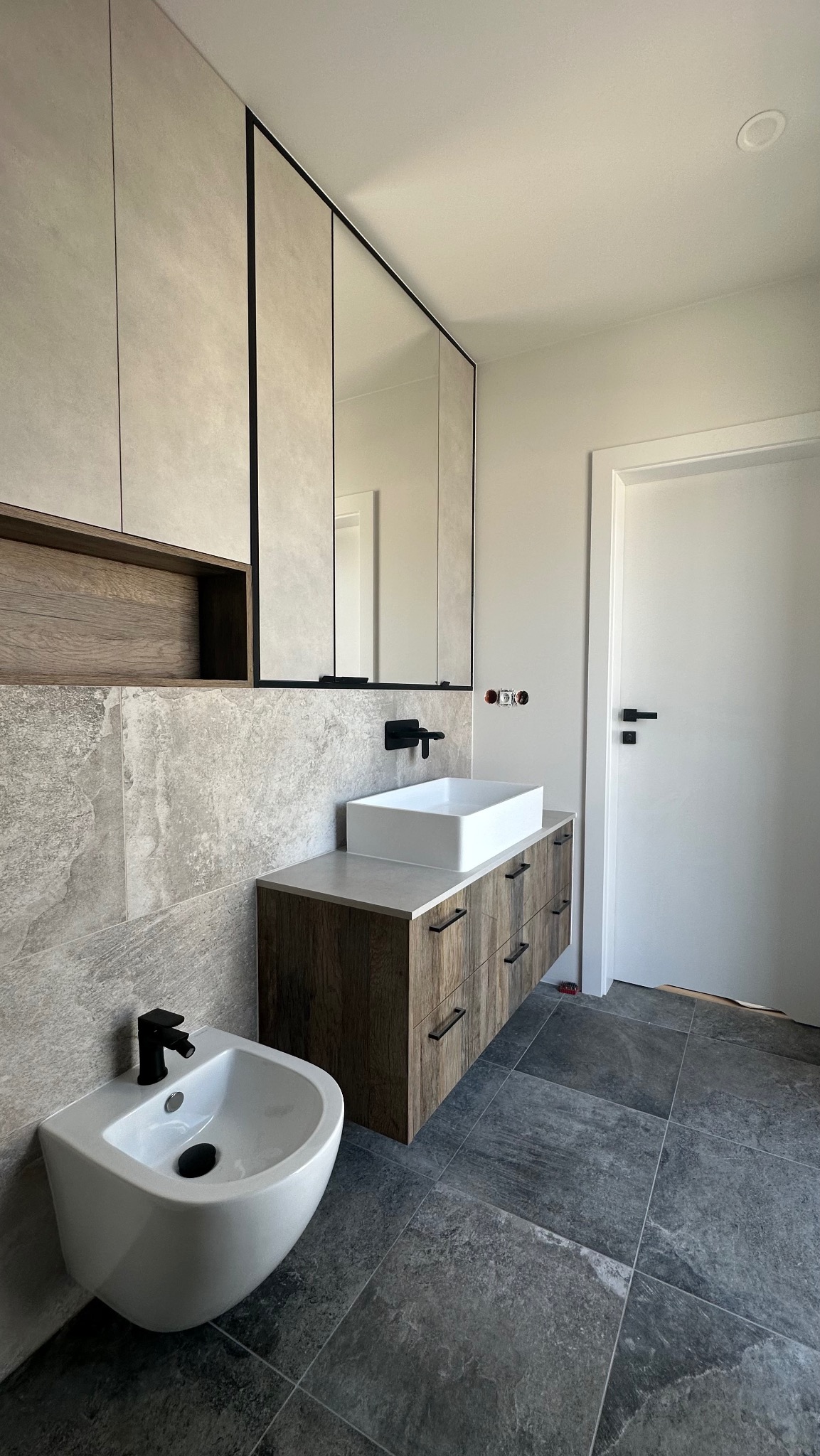 Nowoczesna łazienka: szare płytki, drewniana szafka z umywalką nablatową i czarną armaturą, wiszące szafki z lustrami. Minimalistyczny design.