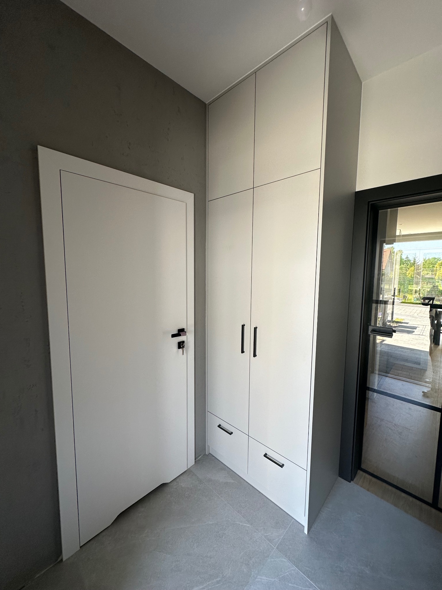 Białe drzwi z czarną klamką sąsiadują z wysoką, jasną szafą wnękową z ciemnymi uchwytami na tle szarej ściany i podłogi z płytek. W tle widoczne przeszklone drzwi.