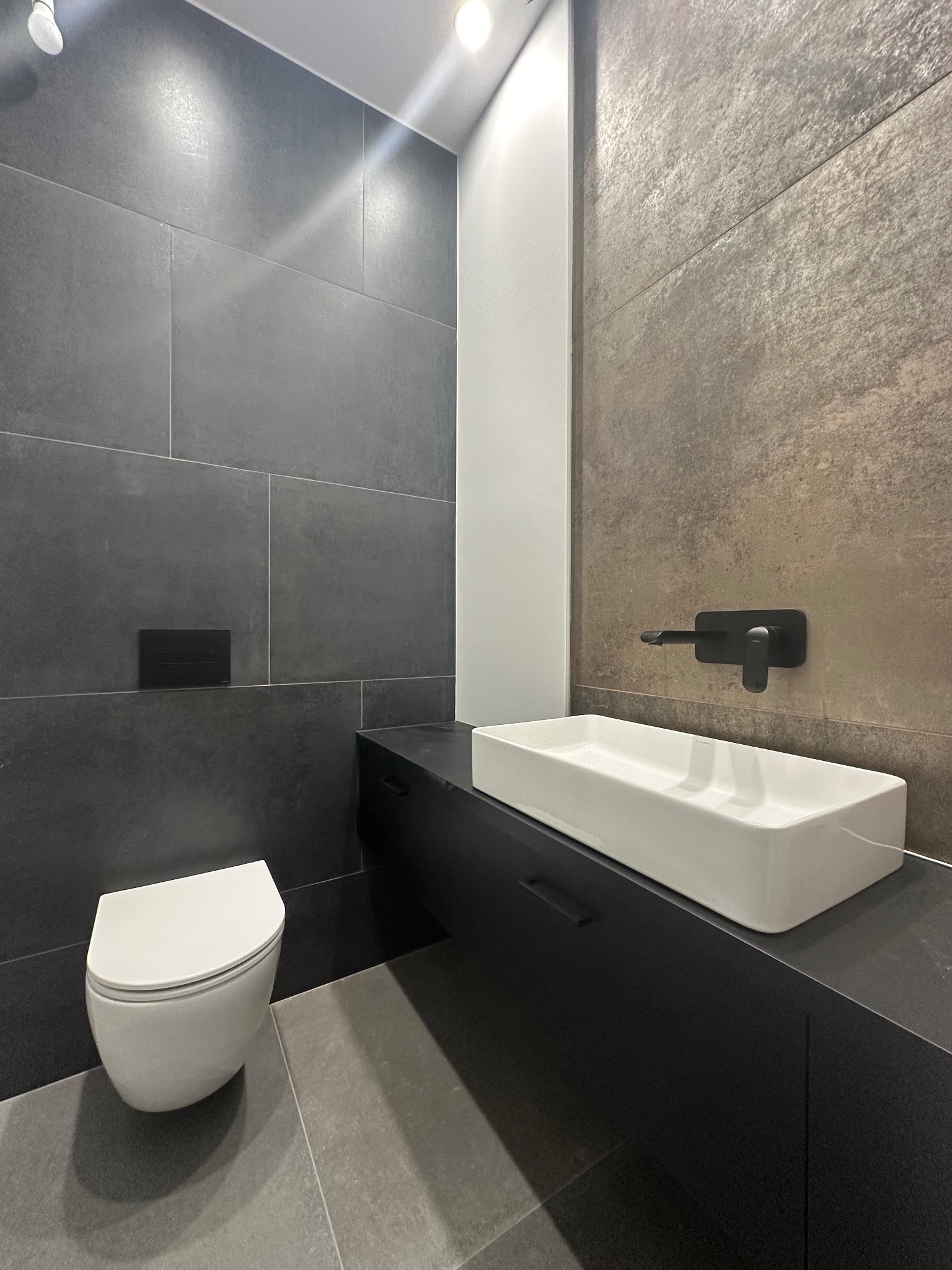 Nowoczesna łazienka z szarą i brązową glazurą, białą umywalką na czarnej szafce i podwieszaną toaletą. Minimalistyczny design, oświetlenie punktowe.