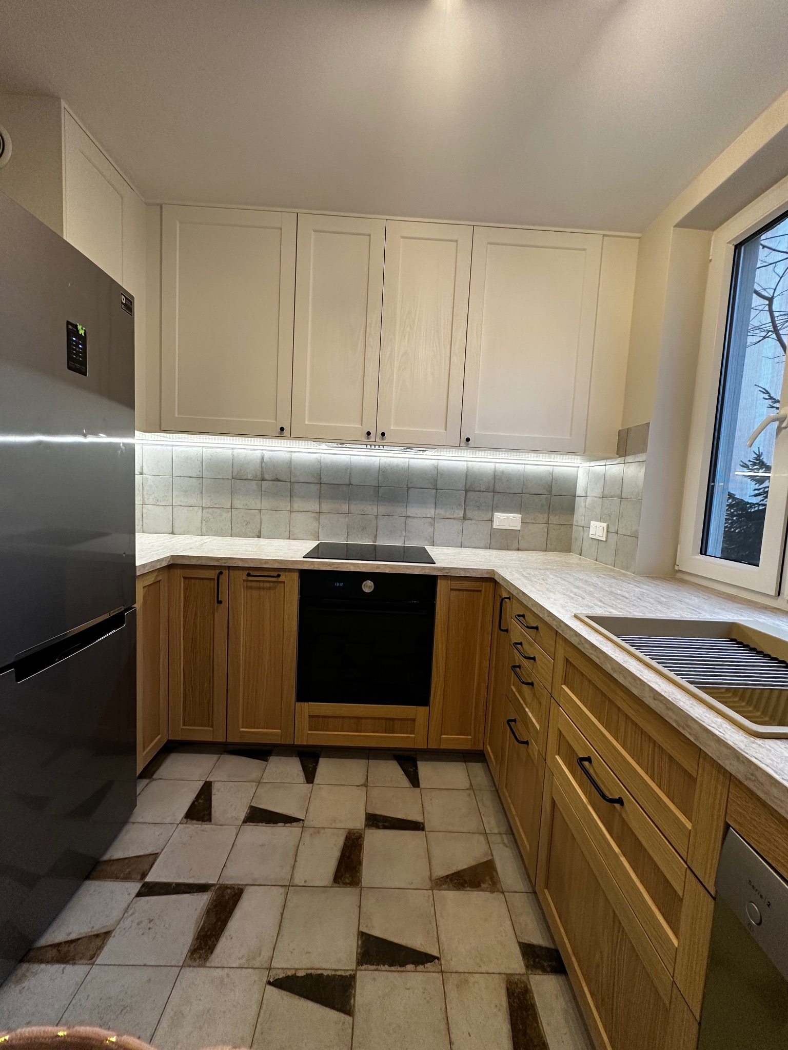 Jasna kuchnia z drewnianymi szafkami i mozaiką. Uwagę przykuwają płytki podłogowe w geometryczne wzory i oświetlenie LED pod szafkami wiszącymi.