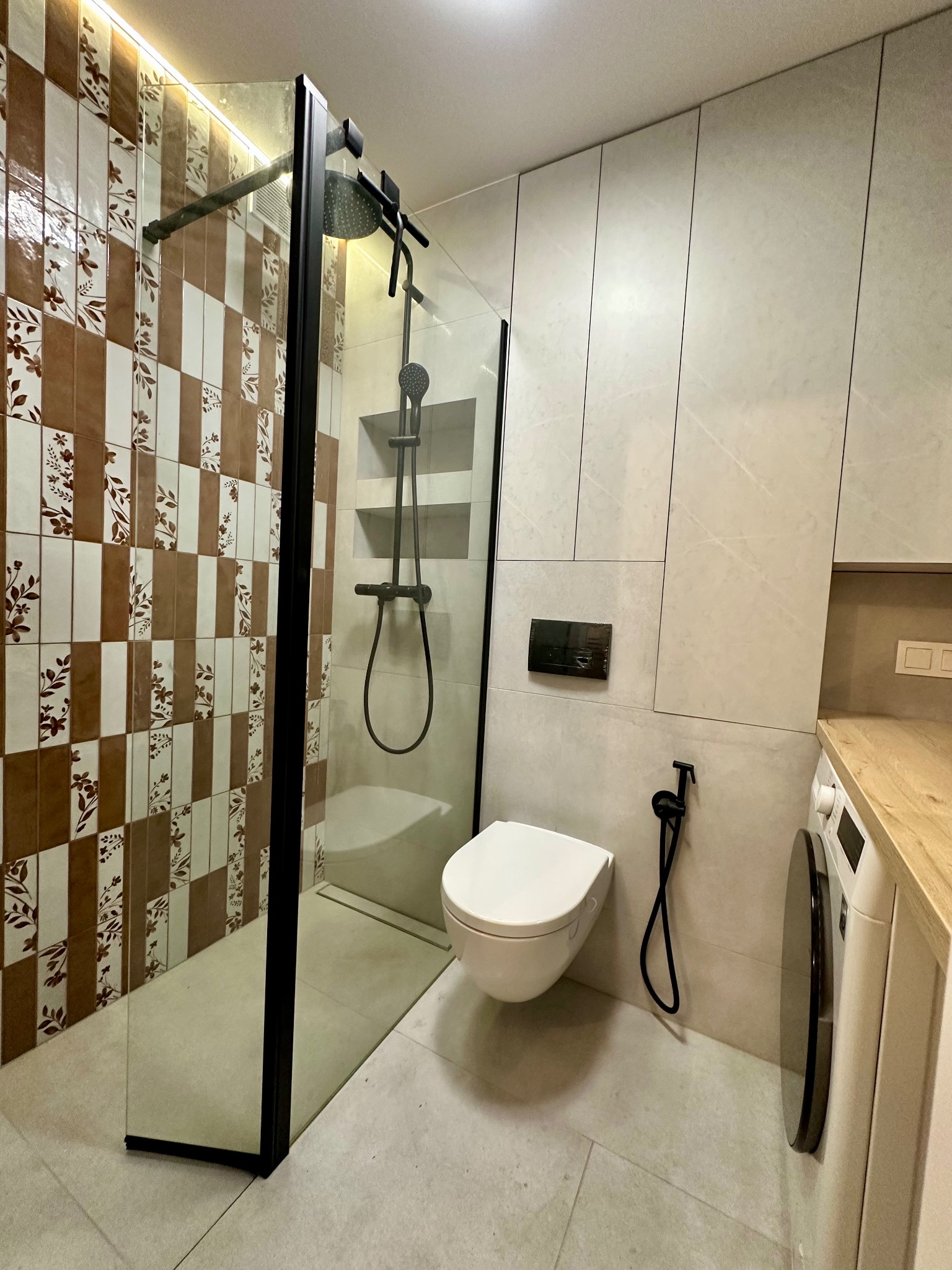 Nowoczesna łazienka w Warszawie z kabiną typu walk-in z czarnymi profilami i dekoracyjnymi płytkami, wisząca toaleta i pralka pod drewnianym blatem.