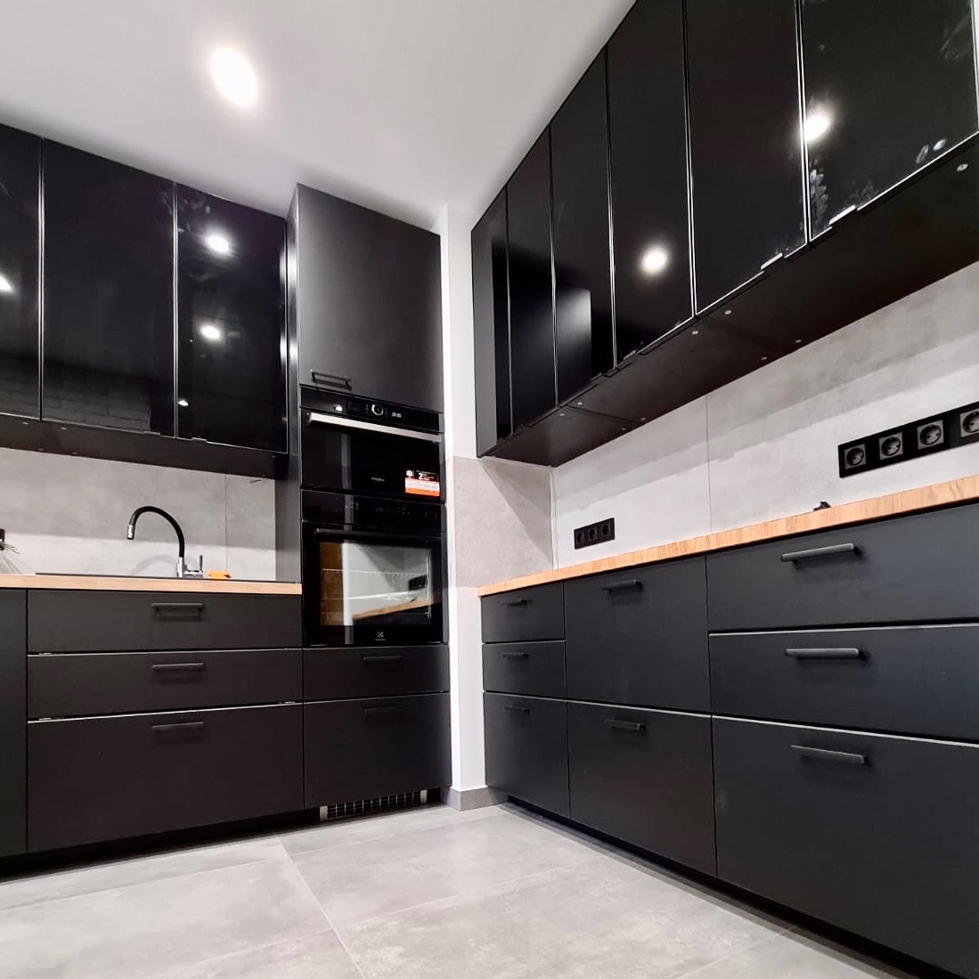 Nowoczesna, czarna kuchnia z drewnianym blatem i błyszczącymi szafkami. Widoczne gniazdka elektryczne na ścianie i piekarnik w zabudowie.