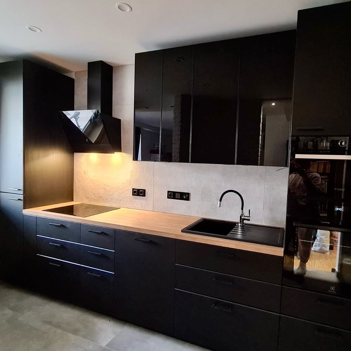 Nowoczesna kuchnia w czerni: matowe fronty szafek dolnych, błyszczące górne. Drewniany blat, szklana płyta grzewcza, czarny zlew i bateria. Minimalistyczny design.