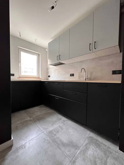 Nowoczesna kuchnia po remoncie: czarne szafki dolne, szare górne, płytki podłogowe imitujące beton, jasne blaty i okno. Minimalistyczny design, surowe wykończenie.