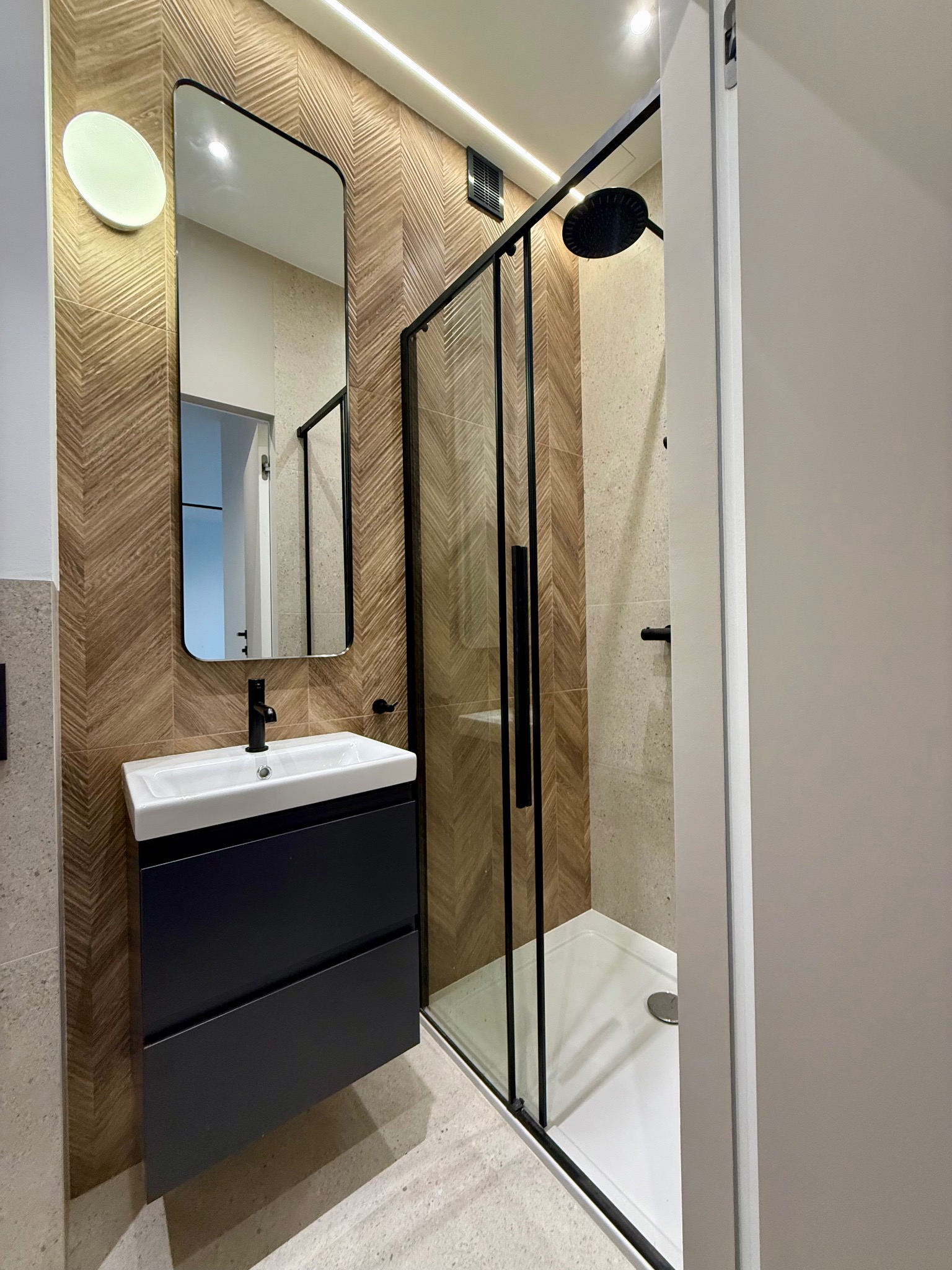 Nowoczesna łazienka z kabiną prysznicową typu walk-in, umywalką na szafce i dużym lustrem. Uwagę zwracają płytki imitujące drewno ułożone w jodełkę na ścianie.