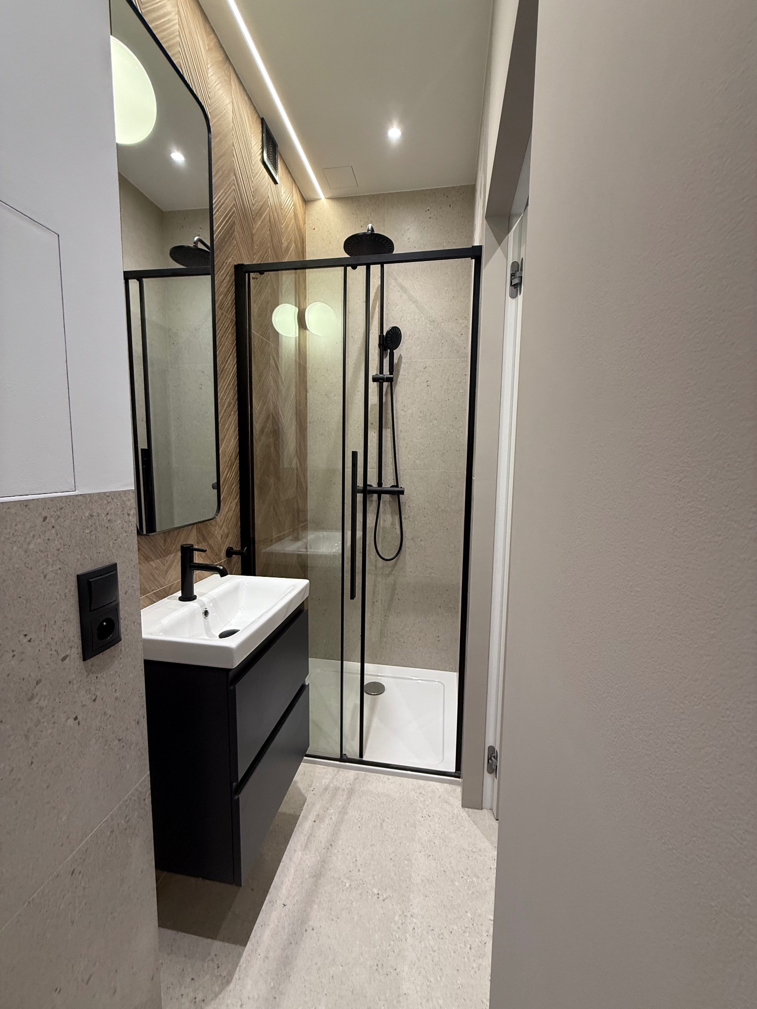 Elegancka łazienka z czarną armaturą i kabiną prysznicową typu walk-in. Umywalka z szafką w kolorze czarnym. Na ścianie lustro z czarną ramą.