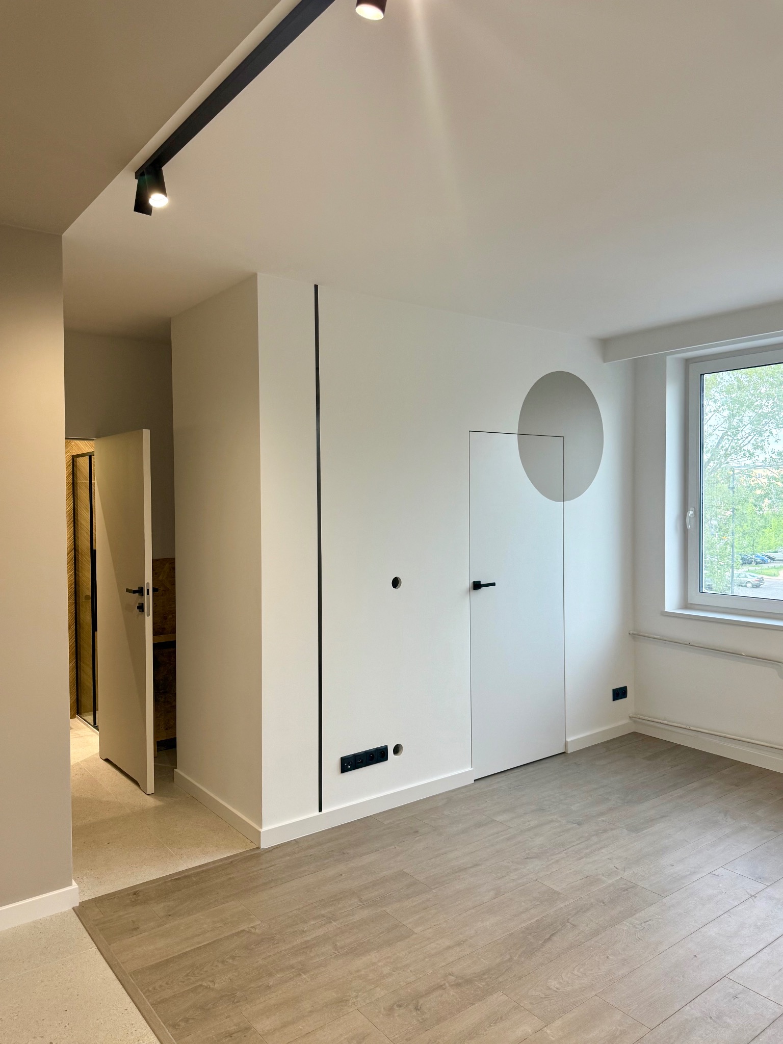 Minimalistyczne wnętrze po remoncie: białe ściany z czarnymi akcentami, ukryte drzwi z szarym okręgiem, panele podłogowe i oświetlenie szynowe.