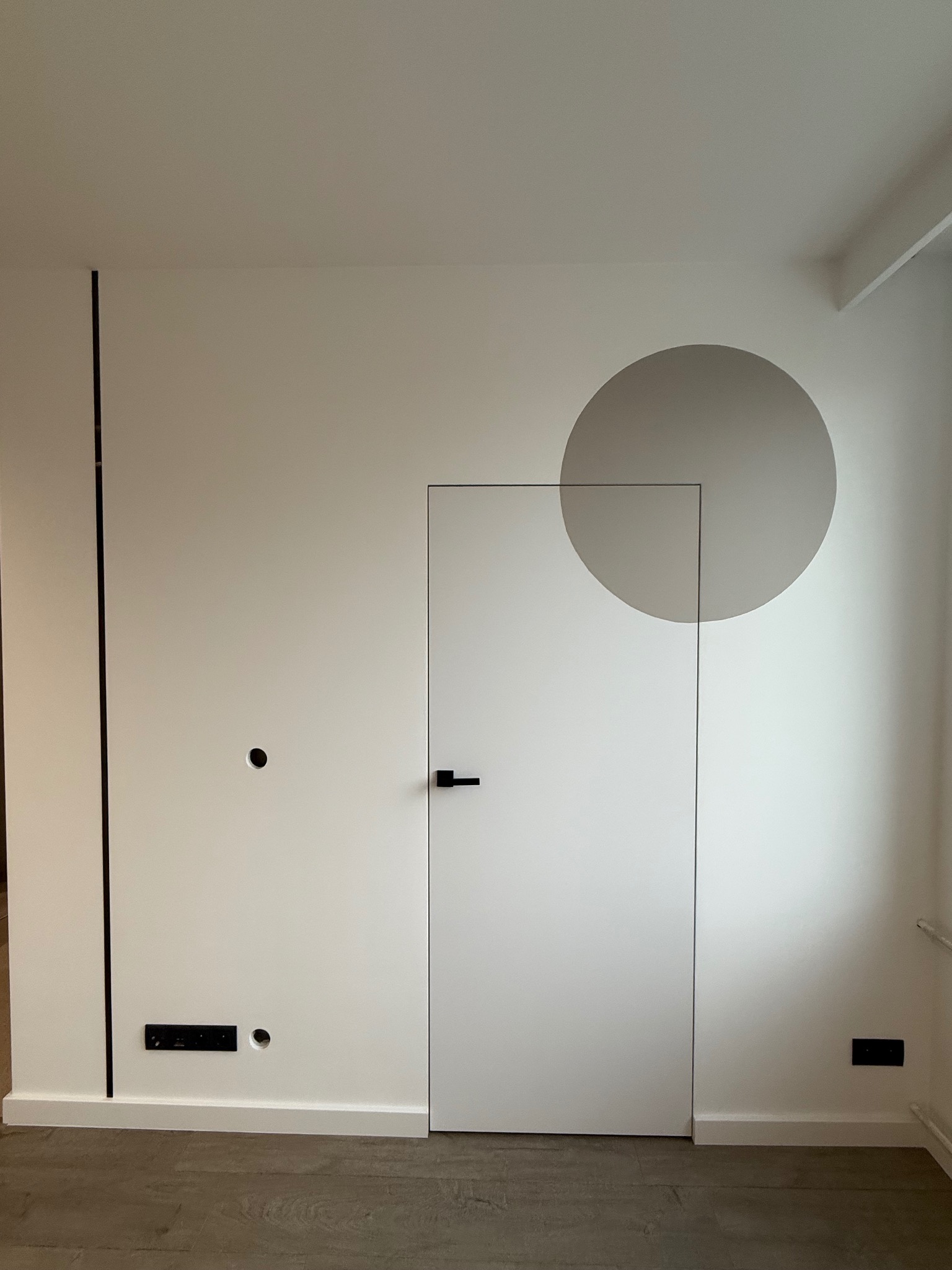 Minimalistyczne wnętrze: biała ściana z ukrytymi drzwiami z czarną klamką, szary okrąg na ścianie, drewniana podłoga, czarne elementy dekoracyjne.