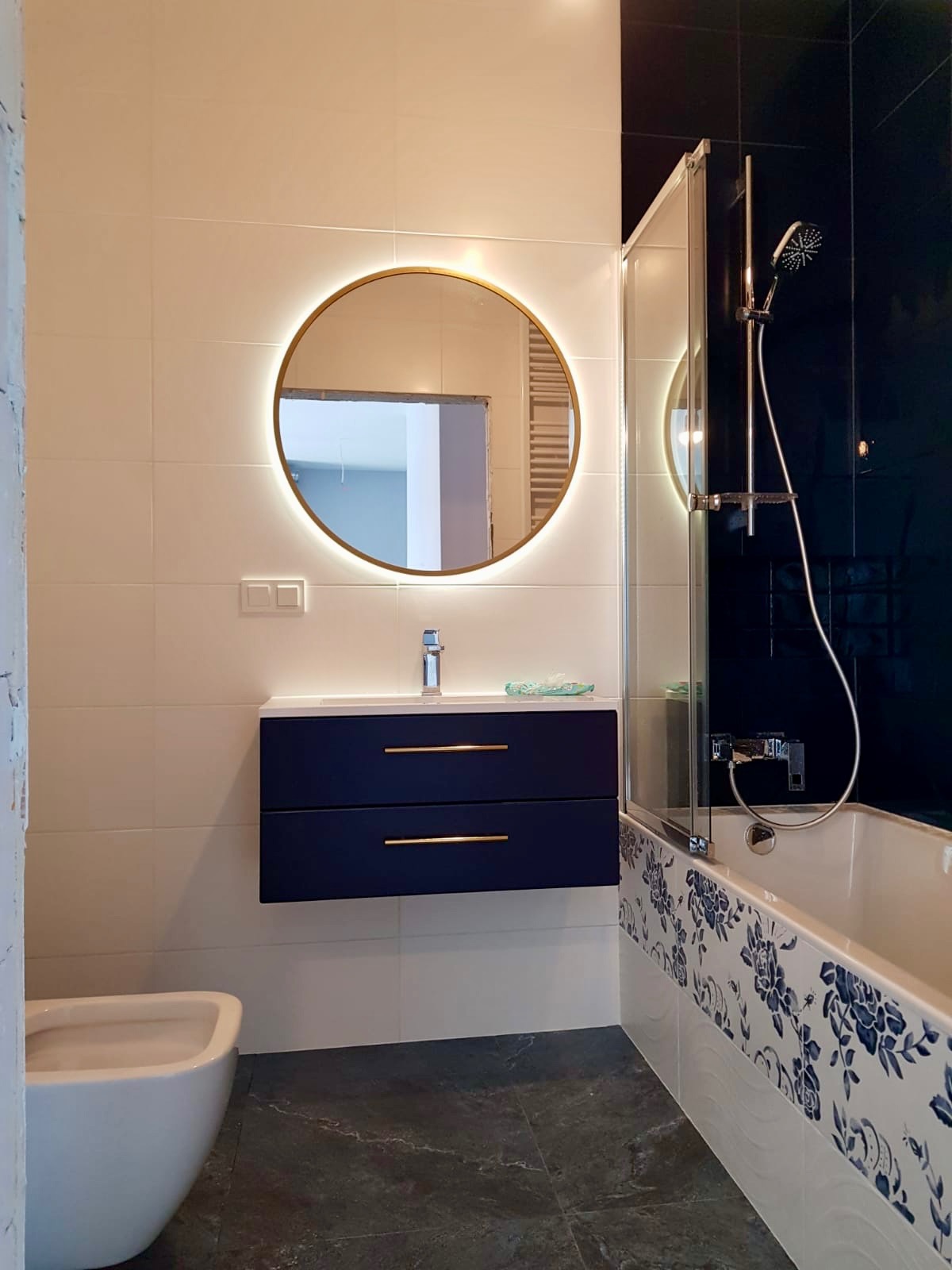 Nowoczesna łazienka po remoncie: białe płytki, szafka z umywalką w kolorze granatowym, okrągłe lustro z podświetleniem, wanna z parawanem i dekoracyjnymi płytkami.