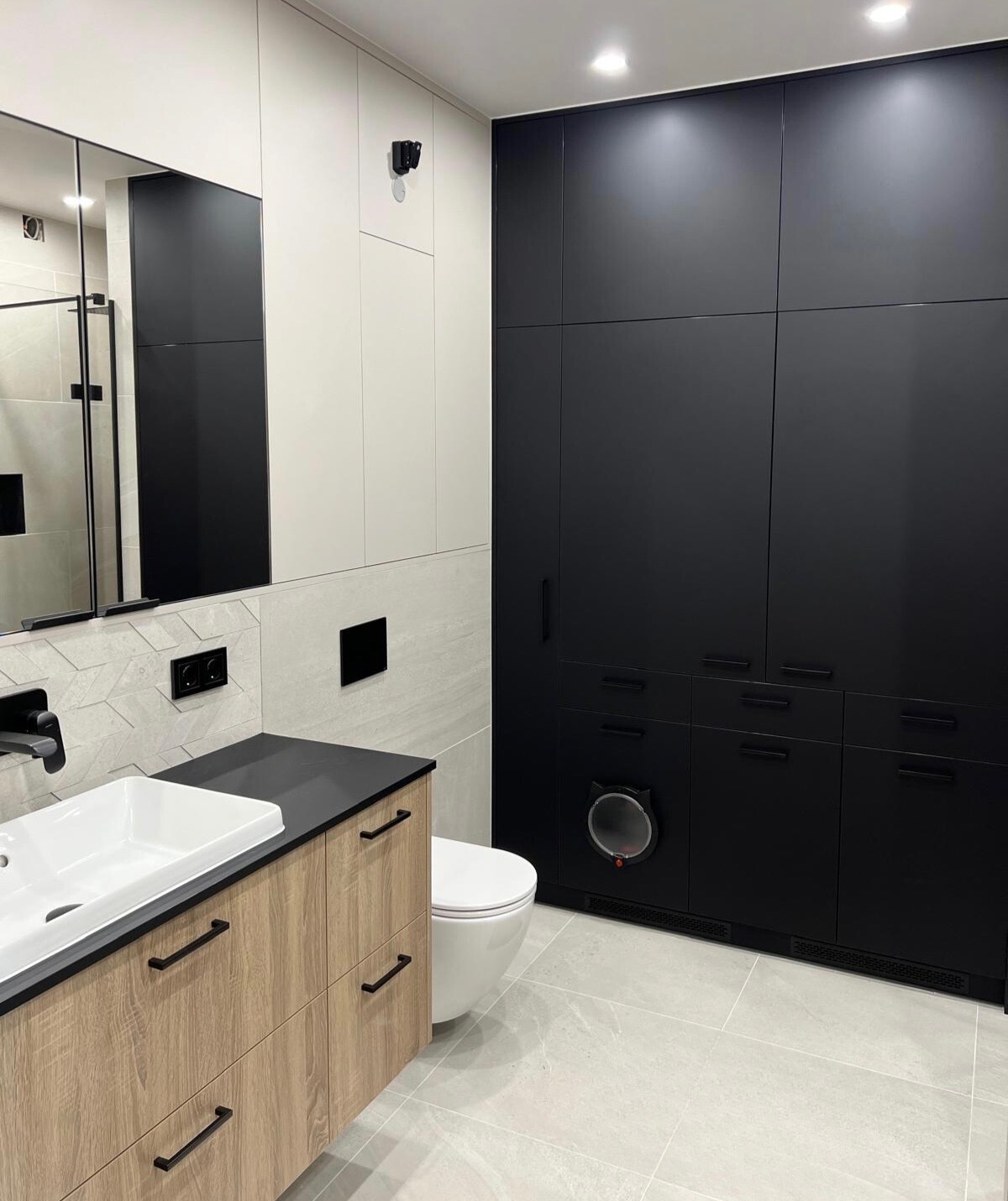 Nowoczesna łazienka po remoncie: jasne płytki, drewnopodobna szafka z umywalką, czarne szafy w zabudowie, toaleta. Nietypowe drzwiczki dla kota w szafce.