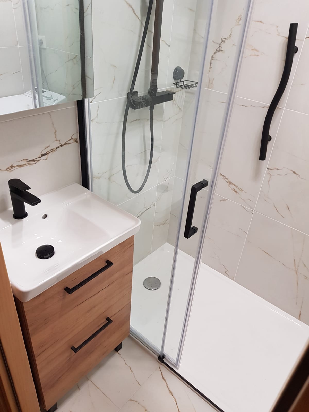 Nowoczesna łazienka po remoncie: kabina prysznicowa z czarną armaturą, umywalka z szafką w odcieniach drewna, marmurowe płytki. Minimalistyczny design, eleganckie wykończenie.