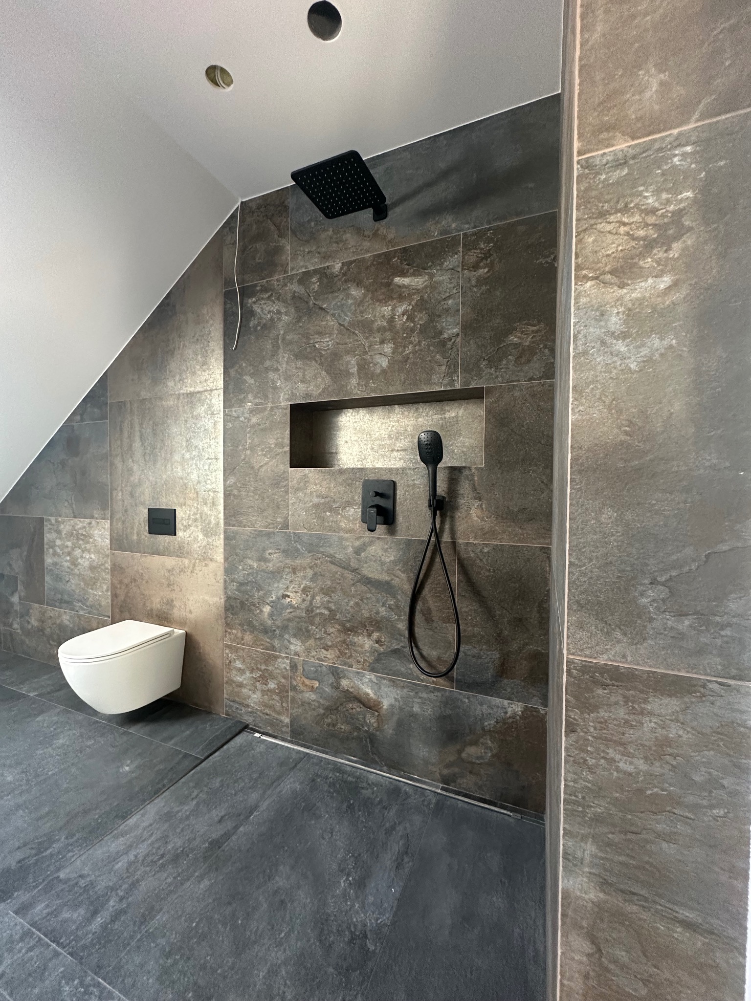 Nowoczesna łazienka na poddaszu z płytkami imitującymi kamień, czarną armaturą i podwieszaną toaletą. Widoczne skosy sufitu i punktowe oświetlenie.