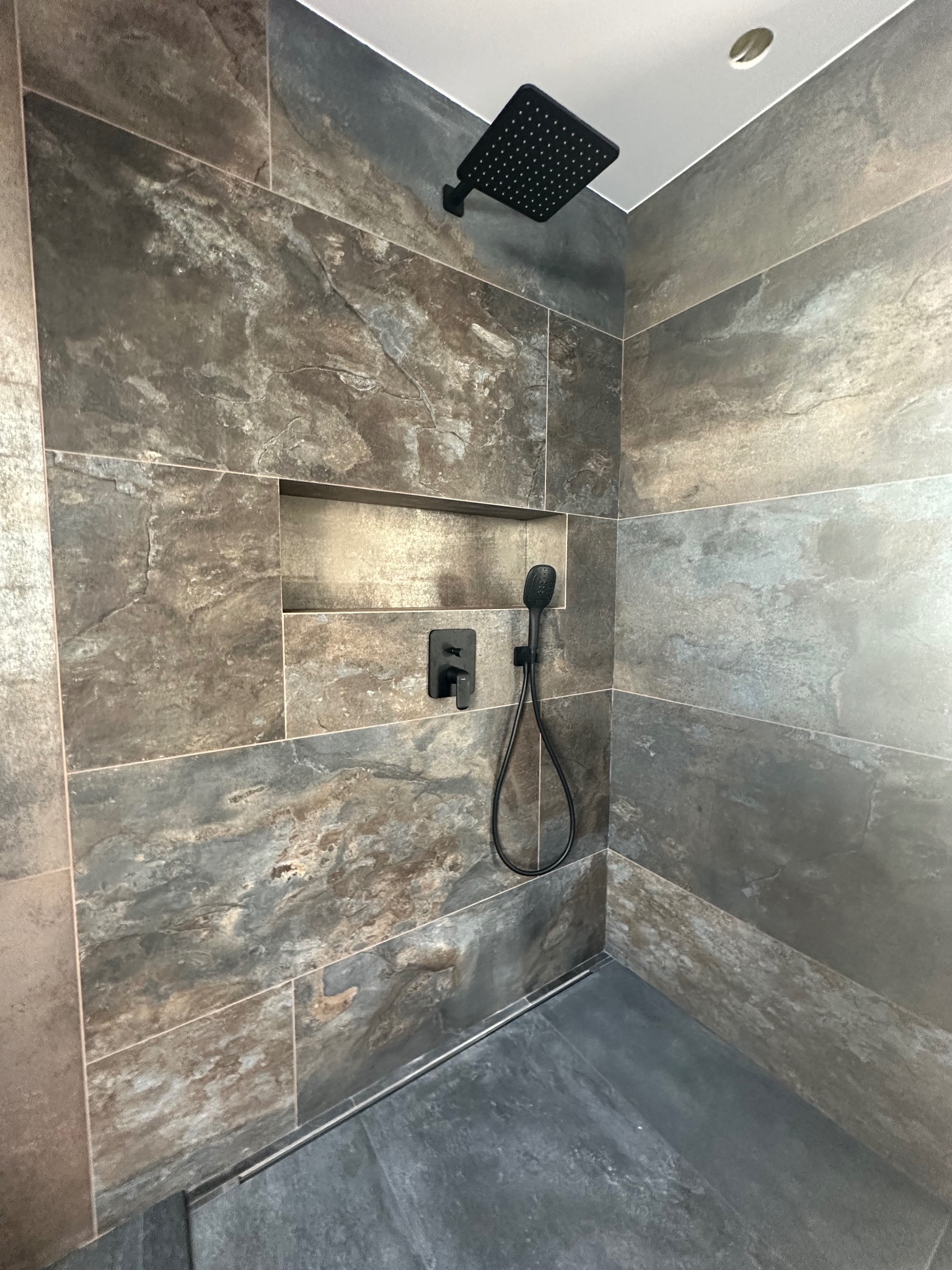 Nowoczesna kabina prysznicowa z ciemnymi, kamiennymi płytkami, wnęką na kosmetyki i czarną armaturą. Minimalistyczny design, szara podłoga, punktowe oświetlenie.