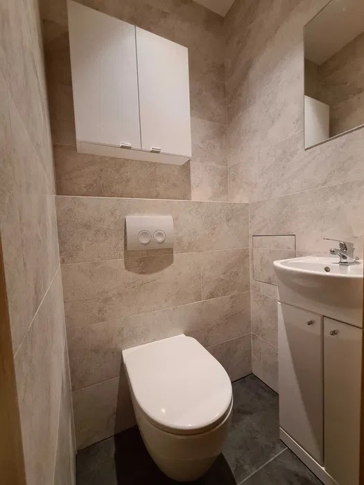 Wykończona łazienka z beżowymi płytkami, wiszącą szafką nad toaletą, umywalką z szafką i lustrem. Ciemna podłoga. Nowoczesny design.