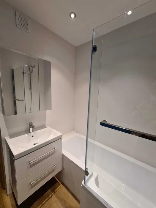 Jasna łazienka po remoncie: biała umywalka z szafką, wanna z parawanem szklanym, lustro z oświetleniem. Minimalistyczny design, drewnopodobna podłoga.