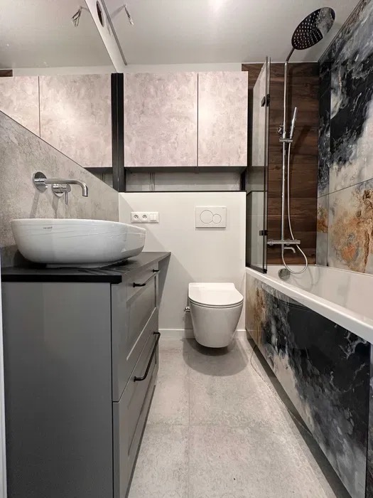 Nowoczesna łazienka po remoncie: szara szafka z umywalką, podwieszana toaleta, wanna obudowana płytkami imitującymi marmur i kabina prysznicowa.