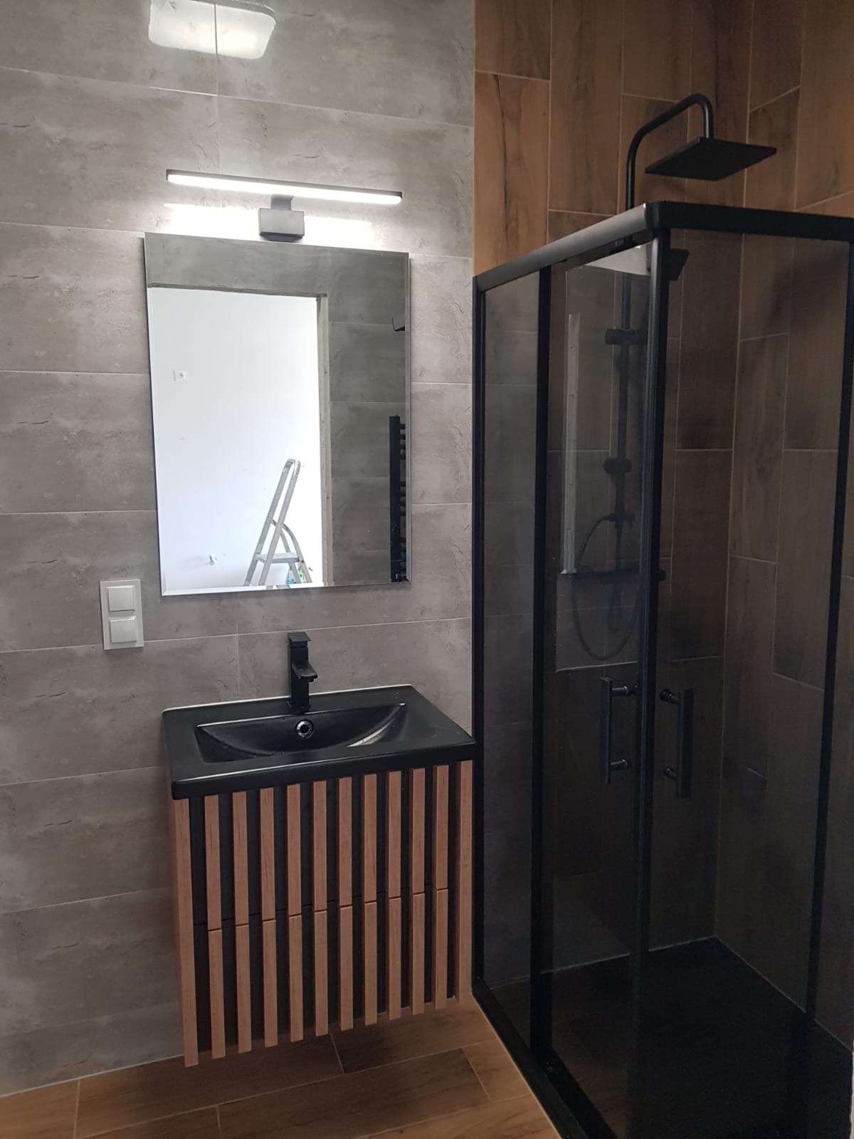 Nowoczesna łazienka z kabiną prysznicową typu walk-in z czarnymi profilami, umywalką z szafką w kolorze drewna i szarymi płytkami na ścianach. Widoczne odbicie drabiny w lustrze.