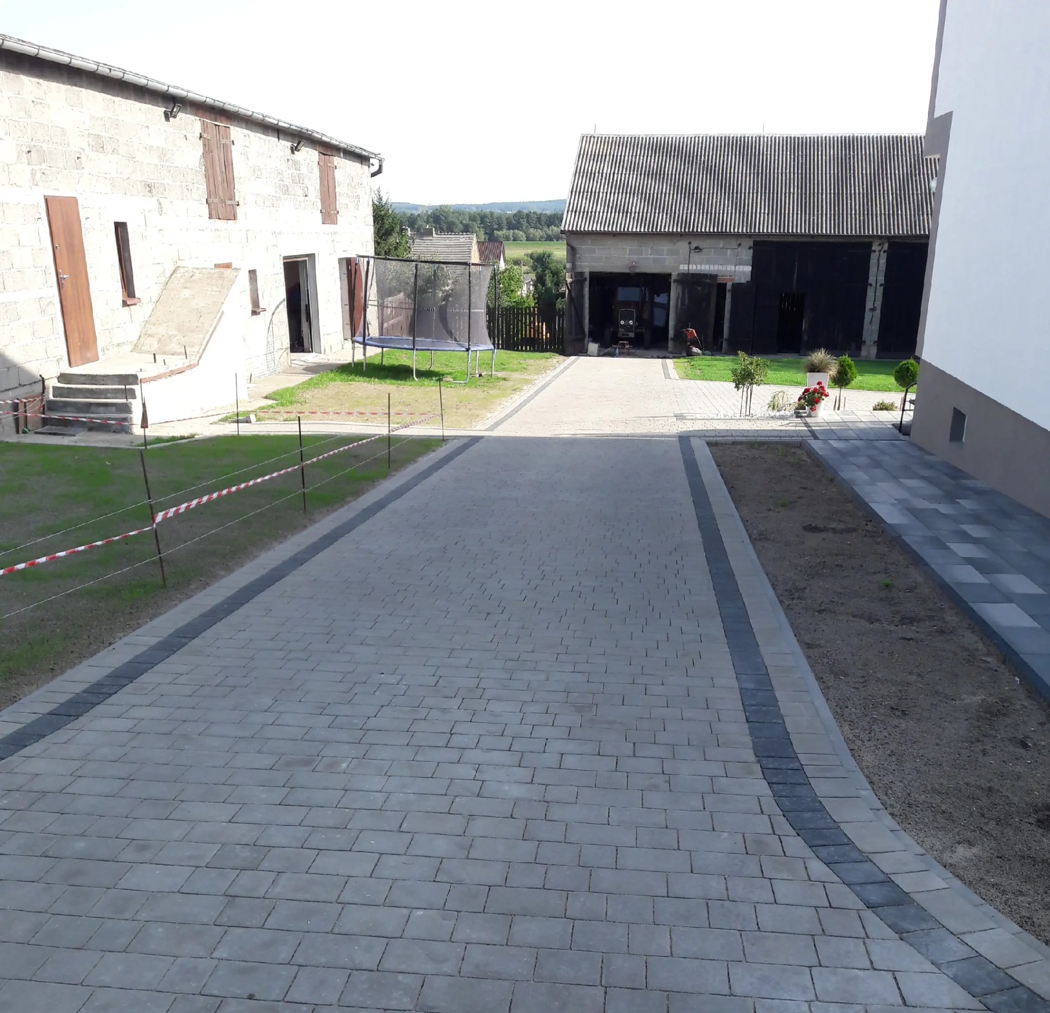 Ułożona kostka brukowa na podjeździe w Sulęczyno. Szara kostka z ciemniejszym obramowaniem. W tle budynki gospodarcze i trampolina.