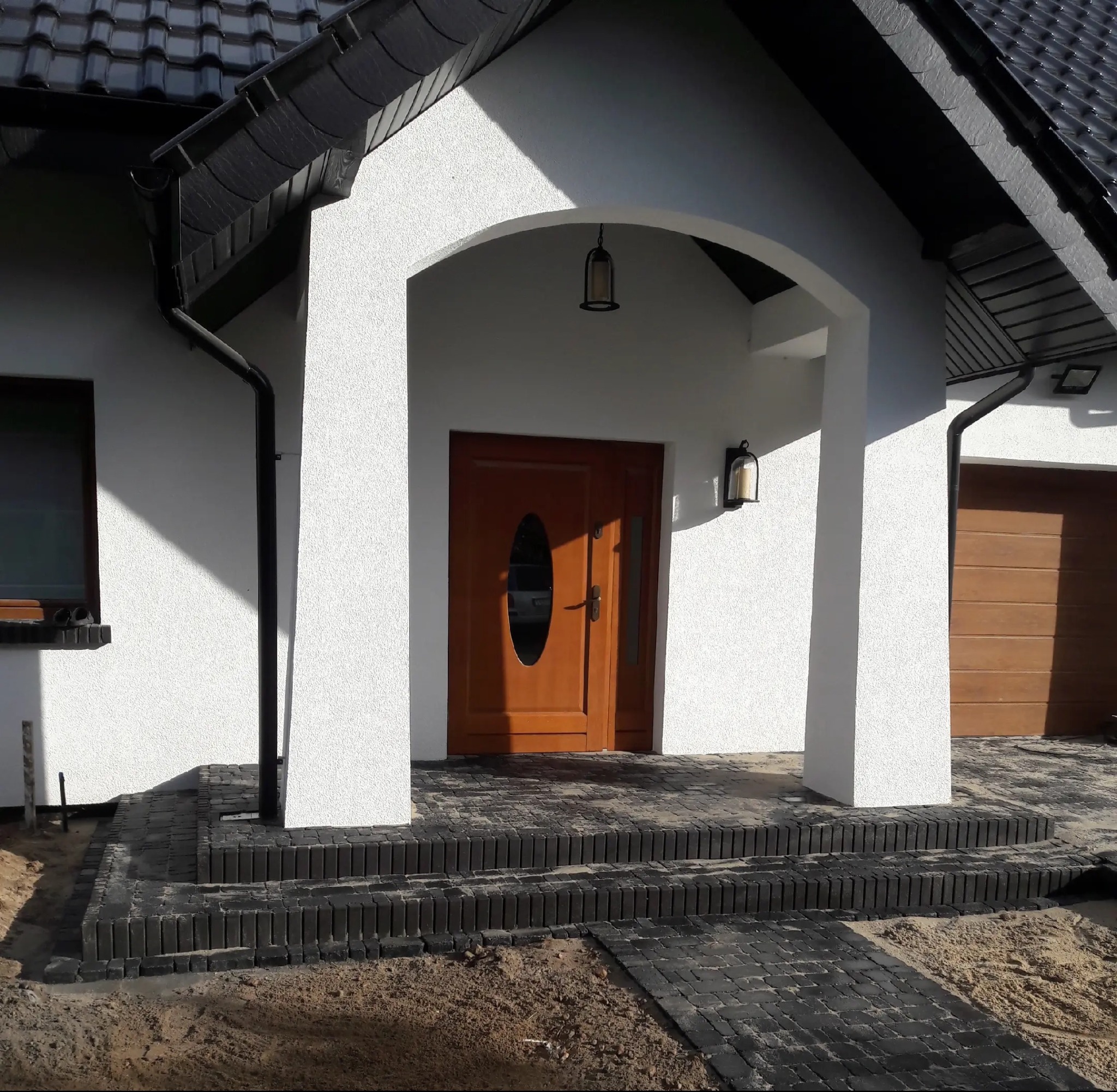 Wejście do domu z ciemnej kostki brukowej, schody i chodnik. Biała elewacja, brązowe drzwi wejściowe z owalnym przeszkleniem. Widoczne oświetlenie zewnętrzne.