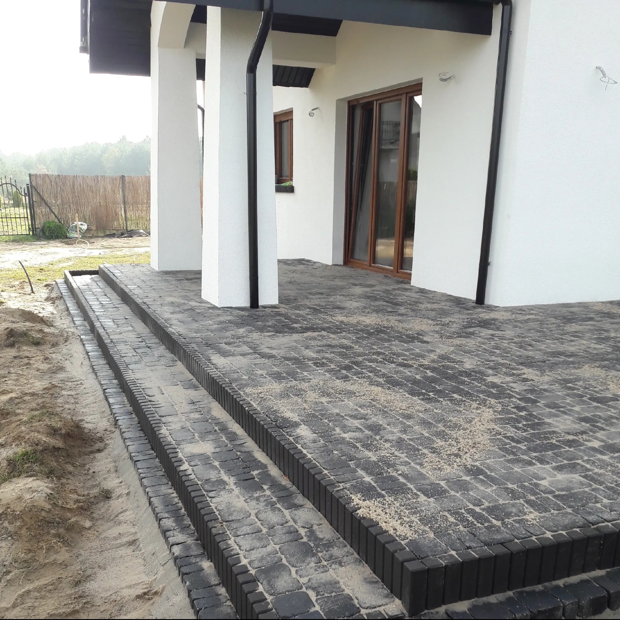 Nowoczesny taras z kostki brukowej w odcieniach szarości, z trzema stopniami prowadzącymi do budynku z białymi kolumnami. Widoczne ślady piasku na kostce.