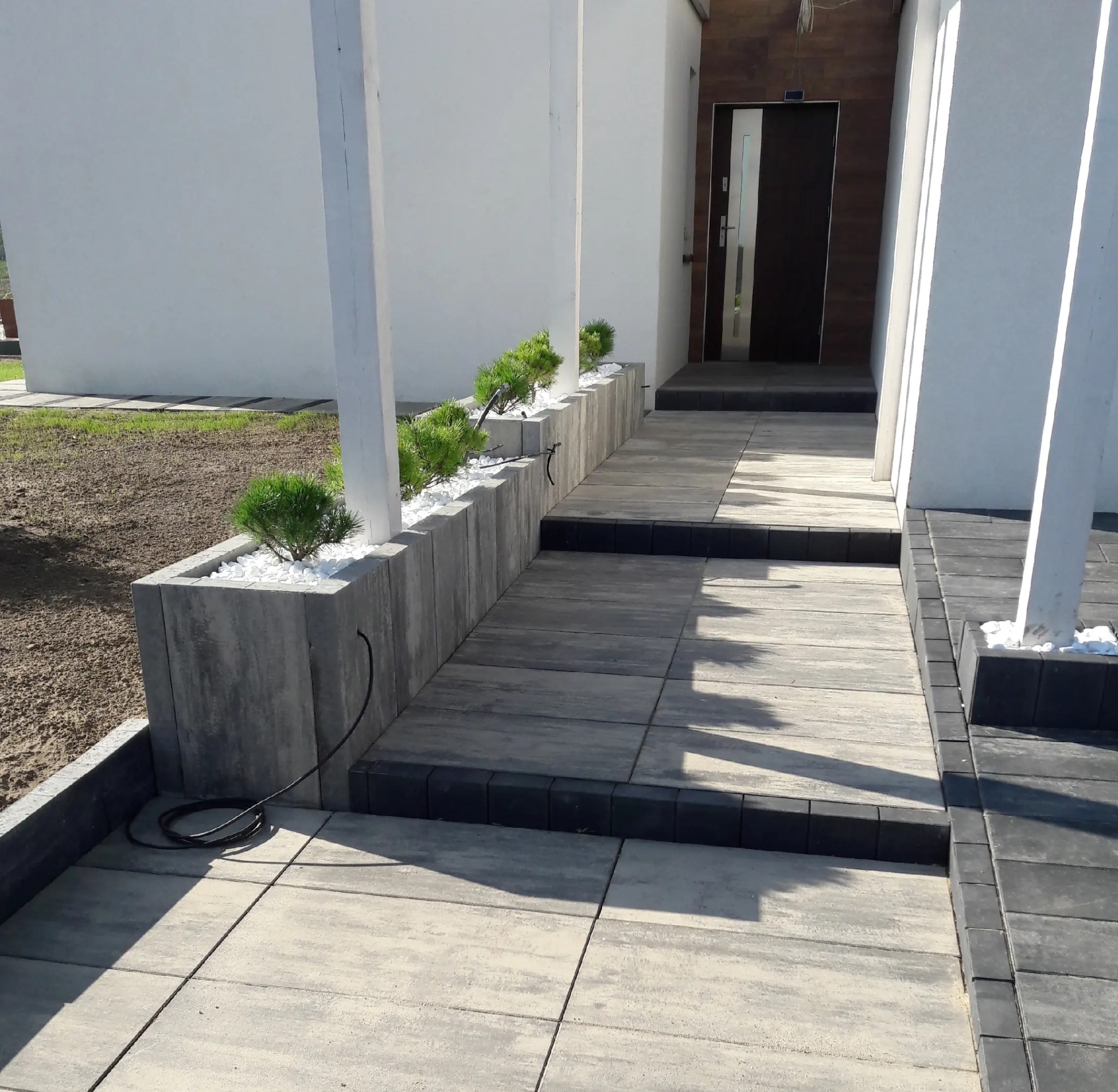 Nowoczesne podejście do domu z kostki brukowej w odcieniach szarości, z elementami dekoracyjnymi w postaci roślin i białych kamieni. Minimalistyczny design.