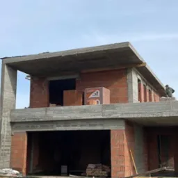 Dwukondygnacyjny budynek w trakcie murowania ścian z czerwonej cegły ceramicznej, z widocznymi elementami żelbetowymi i zapasem materiałów budowlanych.