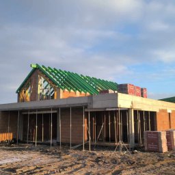 DREAM BUD DOMINIK SOPATA - Budowa domu z keramzytu w stanie surowym otwartym. Widoczne ściany z pustaków, zielona więźba dachowa oraz podpory szalunkowe. Na ścianach ułożone palety z bloczkami.