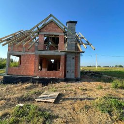 DREAM BUD DOMINIK SOPATA - Wznoszenie dwukondygnacyjnego domu z keramzytu: ściany z pustaków, komin z szarej cegły, drewniana konstrukcja dachu w budowie na tle błękitnego nieba. Paleta na ziemi.