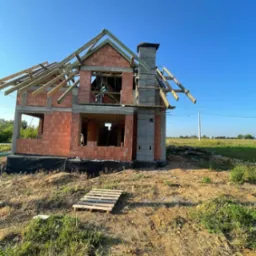 Wznoszenie dwukondygnacyjnego domu z keramzytu: ściany z pustaków, komin z szarej cegły, drewniana konstrukcja dachu w budowie na tle błękitnego nieba. Paleta na ziemi.