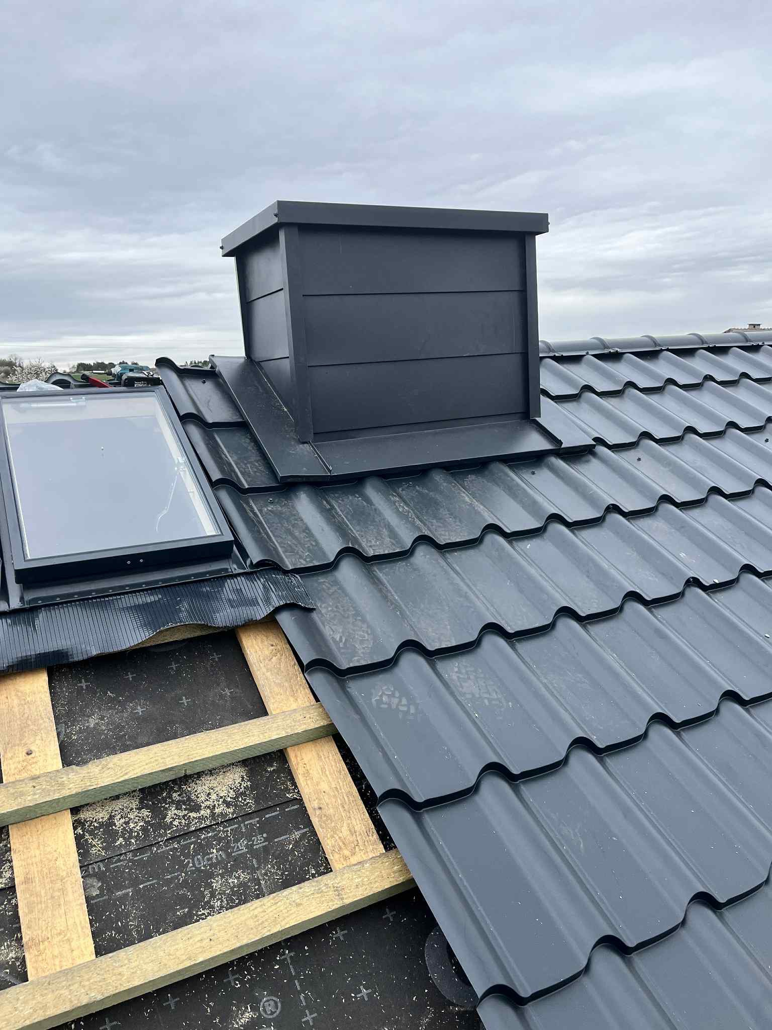 Montaż dachu z blachodachówki z oknem dachowym i kominem w kolorze grafitowym. Widoczne elementy konstrukcji dachu i membrana dachowa.