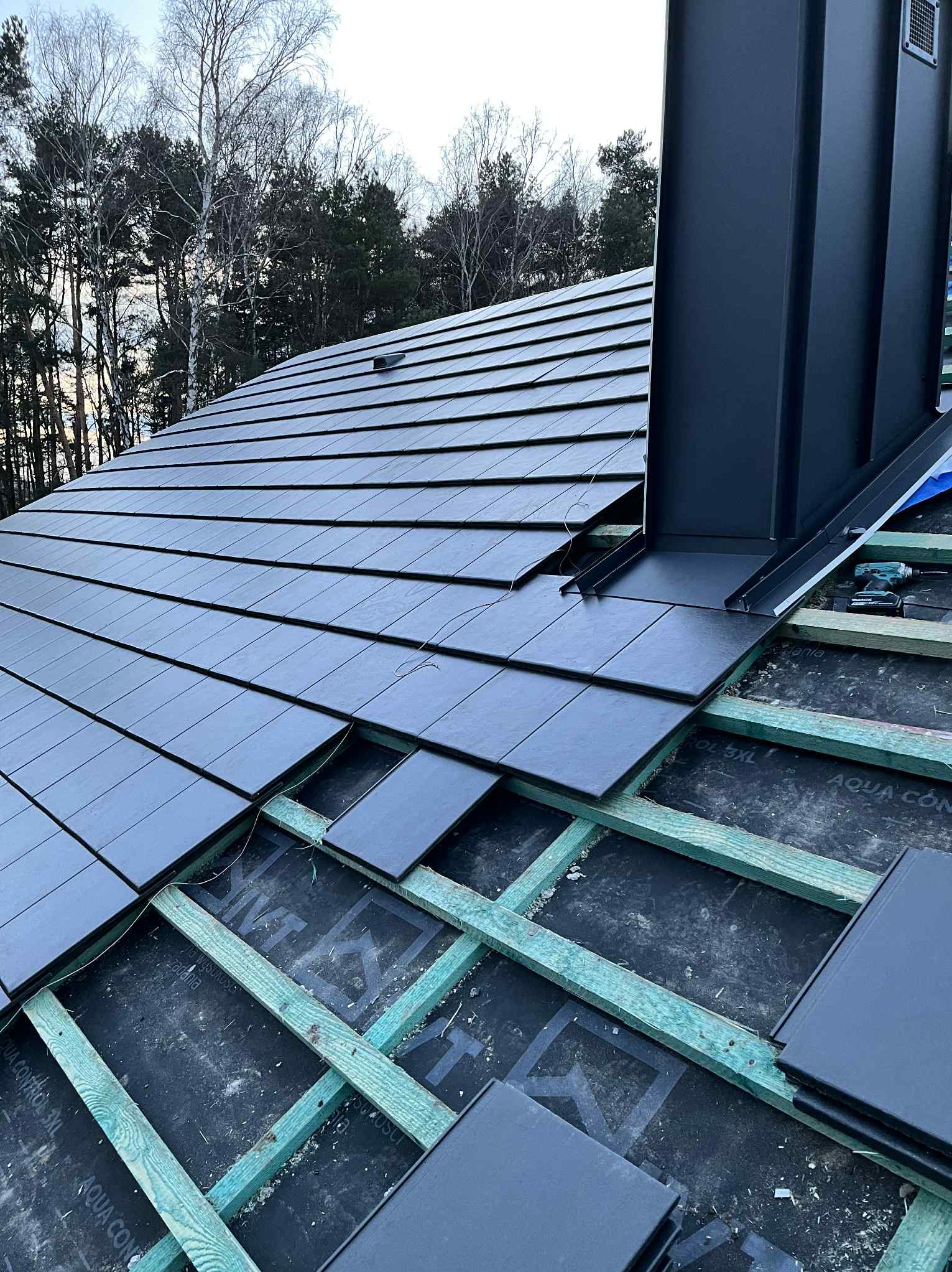 Układanie czarnej dachówki ceramicznej na drewnianej konstrukcji dachu z widocznym kominem i narzędziami. Prace dekarskie w toku, widok z bliska.