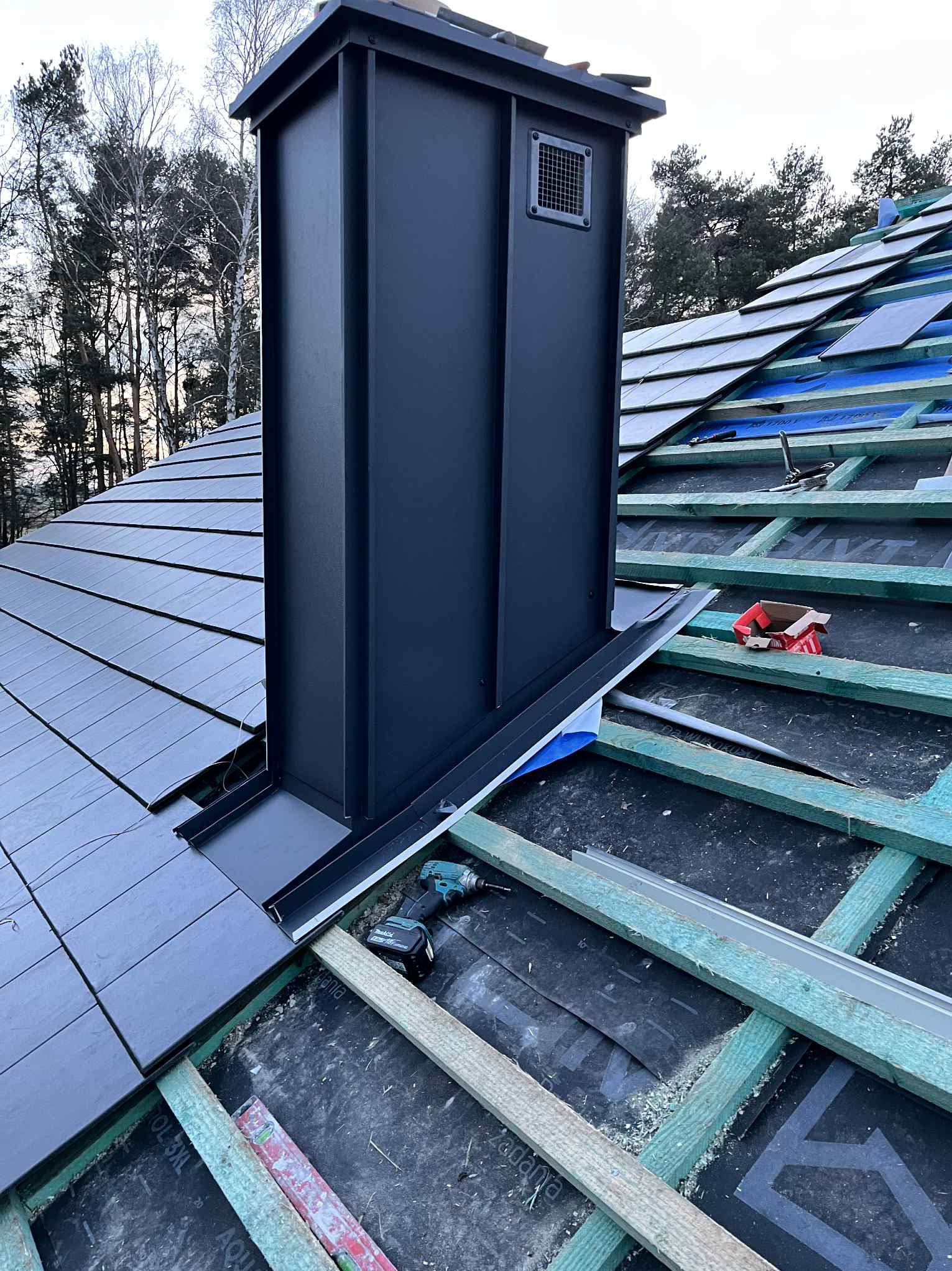 Montaż czarnego komina systemowego na dachu z szarych płytek. Widoczne narzędzia i elementy konstrukcji dachu. Praca w toku, dachówka ceramiczna.