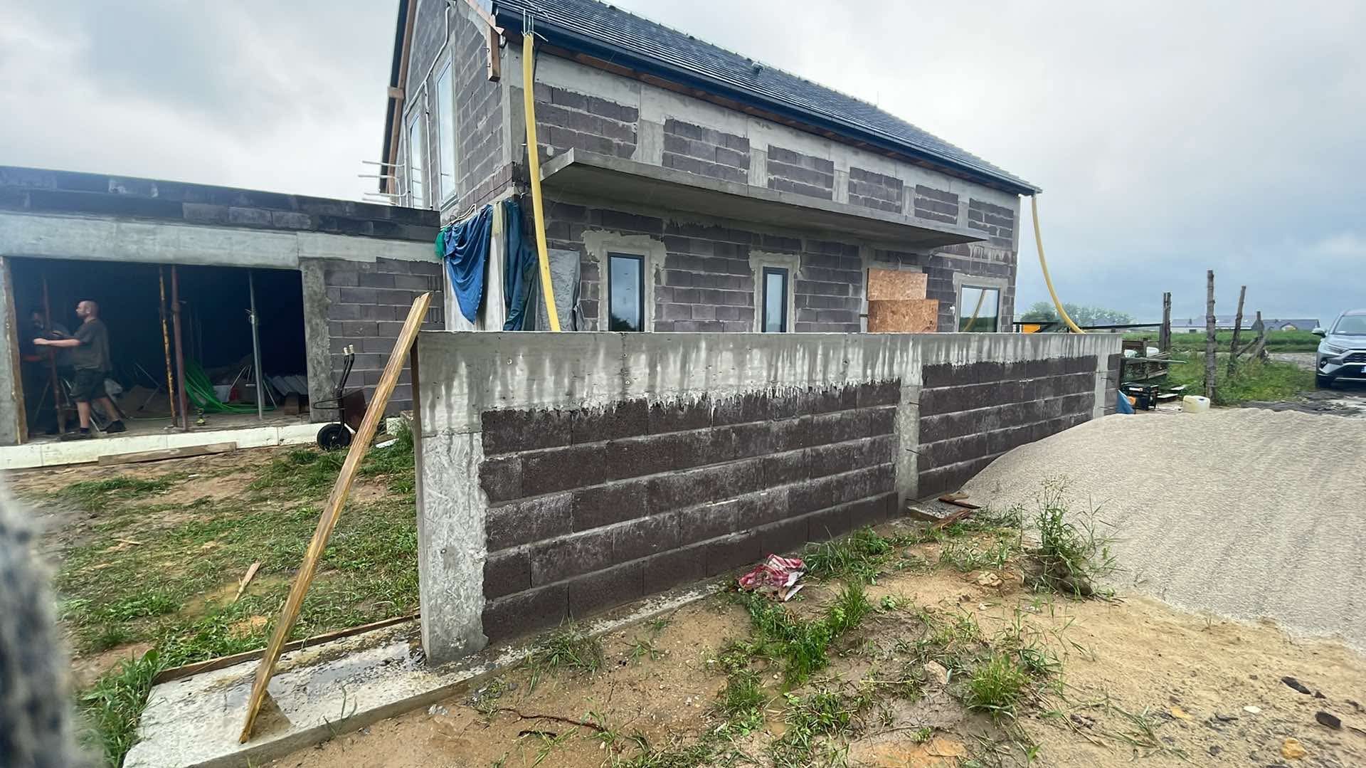 Surowy stan domu z keramzytu w trakcie budowy, z widocznym murem oporowym i stertą piasku na pierwszym planie, na tle pochmurnego nieba.