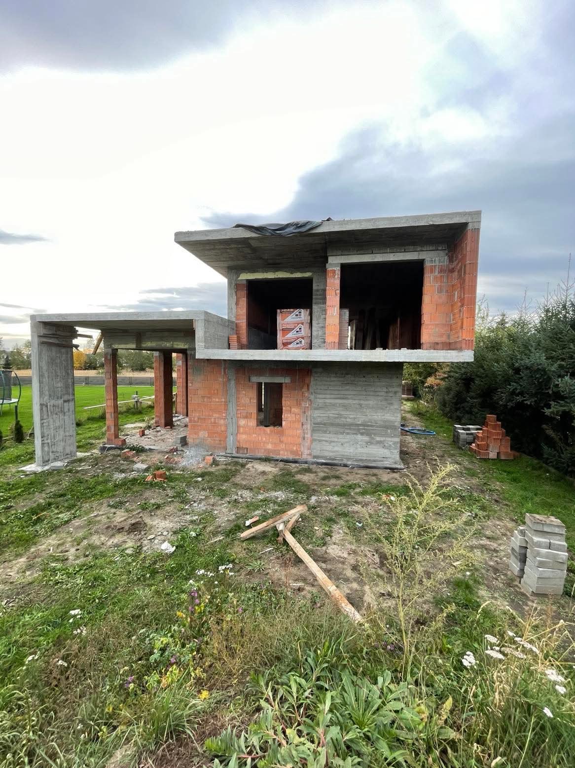 Surowy stan domu murowanego z betonu i cegły na tle pochmurnego nieba. Widoczne niedokończone ściany i konstrukcja dachu.