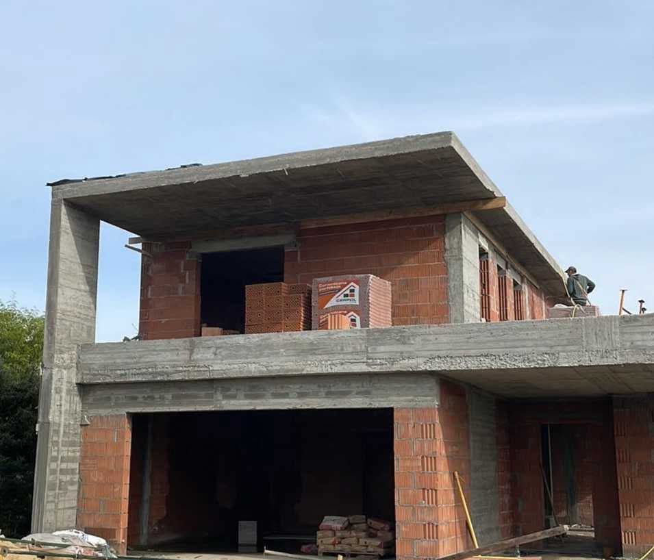 Dwukondygnacyjny budynek w trakcie murowania ścian z czerwonej cegły ceramicznej, z widocznymi elementami żelbetowymi i zapasem materiałów budowlanych.