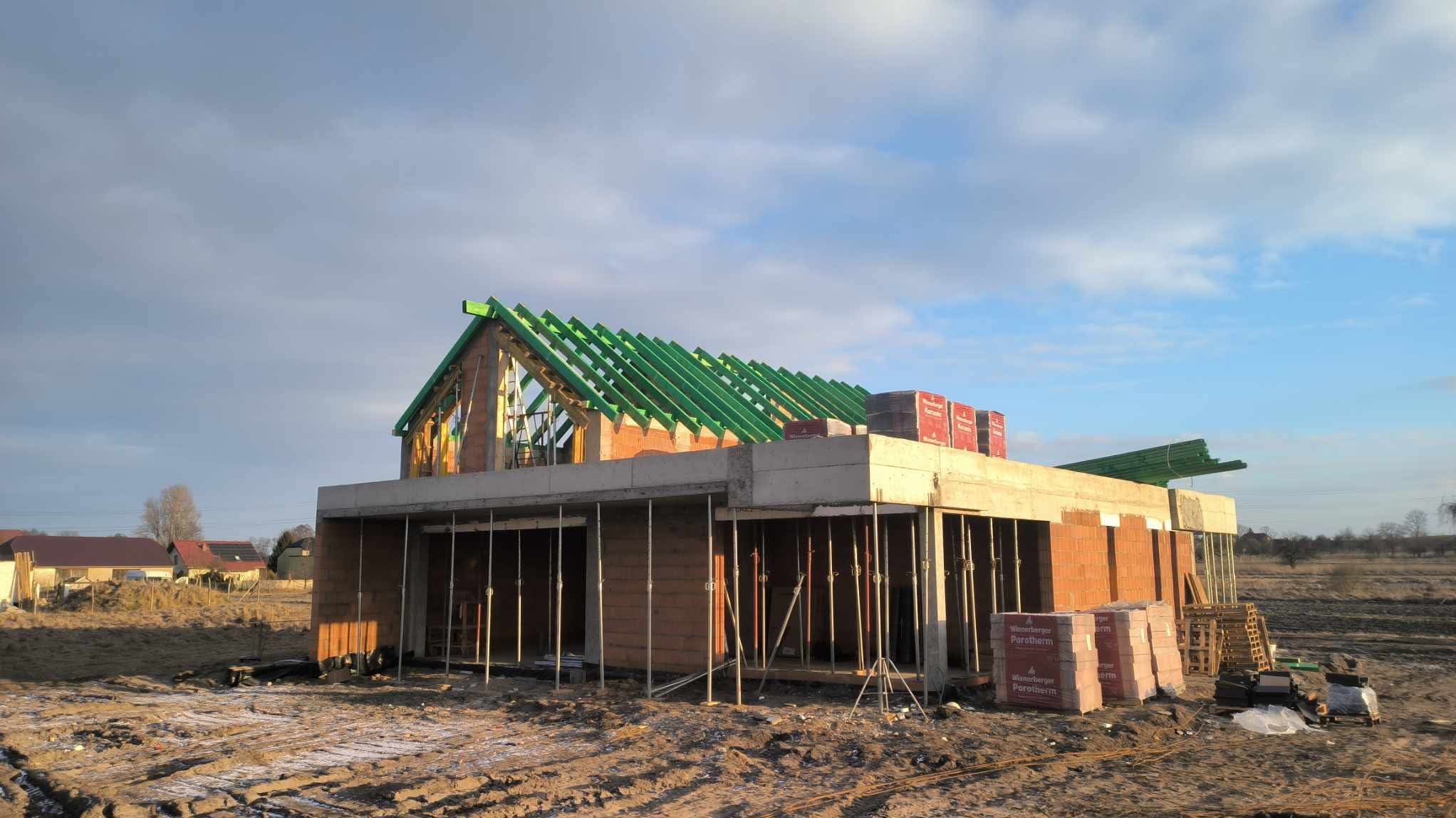 Budowa domu z keramzytu w stanie surowym otwartym. Widoczne ściany z pustaków, zielona więźba dachowa oraz podpory szalunkowe. Na ścianach ułożone palety z bloczkami.
