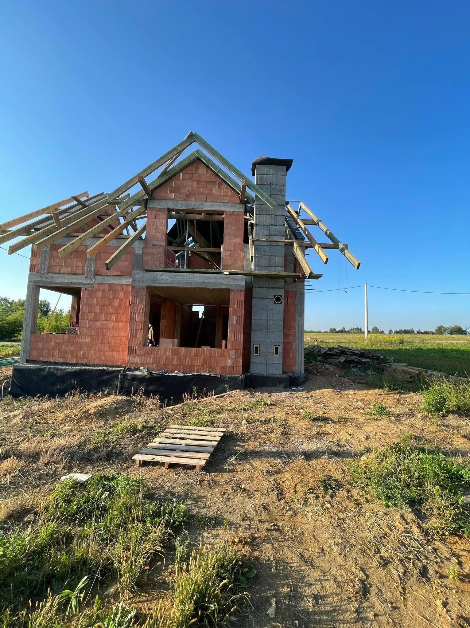 Wznoszenie dwukondygnacyjnego domu z keramzytu: ściany z pustaków, komin z szarej cegły, drewniana konstrukcja dachu w budowie na tle błękitnego nieba. Paleta na ziemi.