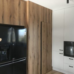 DARCO-USŁUGI MONTAŻOWE ZABIELSKI DARIUSZ - Nowoczesna kuchnia z zabudową drewnopodobną, integrującą czarną lodówkę side-by-side. Białe szafki z piekarnikiem i minimalistycznymi uchwytami dopełniają aranżację.