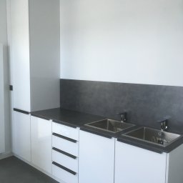 DARCO-USŁUGI MONTAŻOWE ZABIELSKI DARIUSZ - Minimalistyczna, biała zabudowa kuchenna z ciemnym blatem i dwoma zlewozmywakami. Nowoczesne, długie uchwyty podkreślają geometrię szafek.