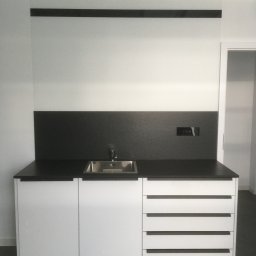 DARCO-USŁUGI MONTAŻOWE ZABIELSKI DARIUSZ - Nowoczesna zabudowa kuchenna z białymi szafkami, czarnym blatem i zlewozmywakiem. Minimalistyczny design, ciemne uchwyty i backsplash.
