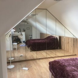 DARCO-USŁUGI MONTAŻOWE ZABIELSKI DARIUSZ - Szafa wnękowa z lustrzanymi drzwiami w pokoju na poddaszu, odbicie wnętrza pokoju. Widoczna część łóżka z bordową narzutą i drewniane wykończenie szafy.